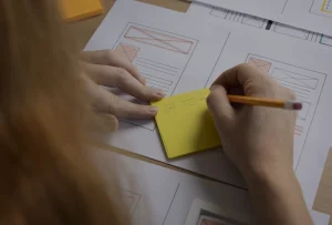 Mão a escrever num post‑it sobre folhas com wireframes, representando o planeamento estratégico de um website.