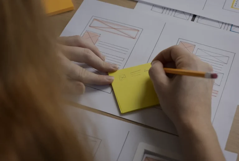 Porque muitos websites falham (mesmo sendo bonitos) Mão a escrever num post‑it sobre folhas com wireframes, representando o planeamento estratégico de um website.