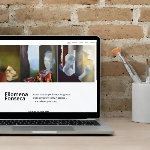 FILOMENA FONSECA ARTES Laptop com o site de Filomena Fonseca aberto, ao lado de pincéis num copo branco sobre mesa de madeira.