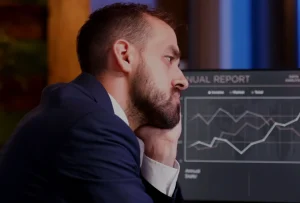 Homem de fato a olhar frustrado para dois monitores com gráficos de queda de performance de um site, num ambiente de escritório.